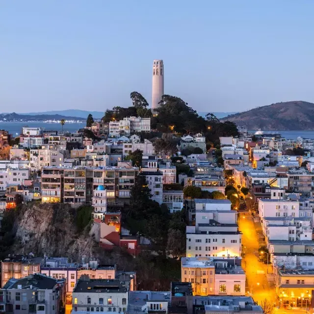 La Torre Coit de San Francisco al anochecer, con calles iluminadas frente a ella y la Bahía de San Francisco detrás.